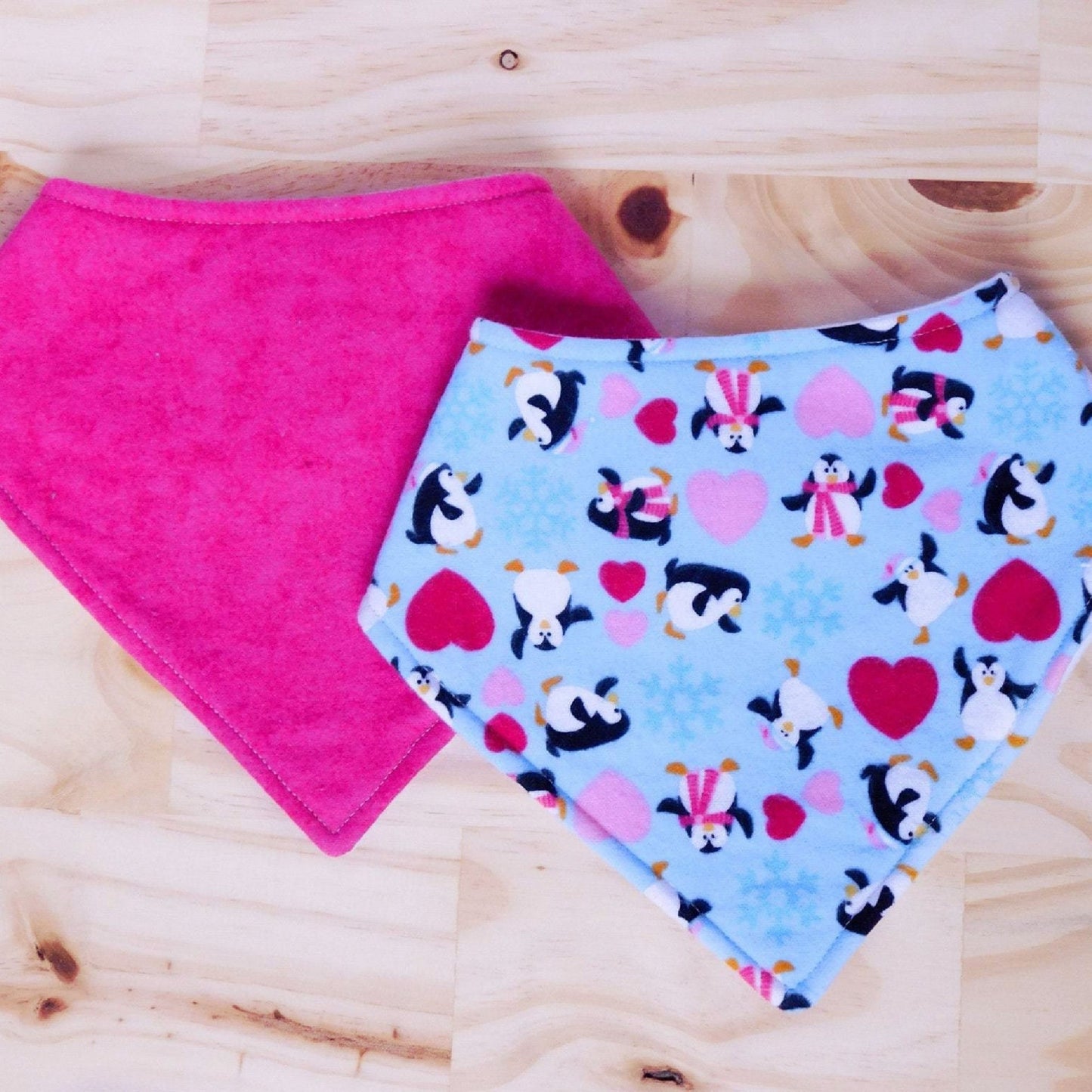 Set of two bandana bibs. Dancing penguins with red and pink hearts on a light blue fabric and solid hot pink bibs