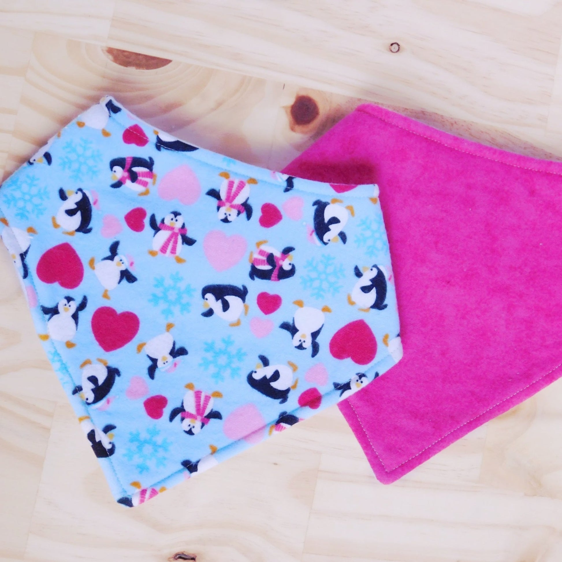 Set of two bandana bibs. Dancing penguins with red and pink hearts on a light blue fabric and solid hot pink bibs